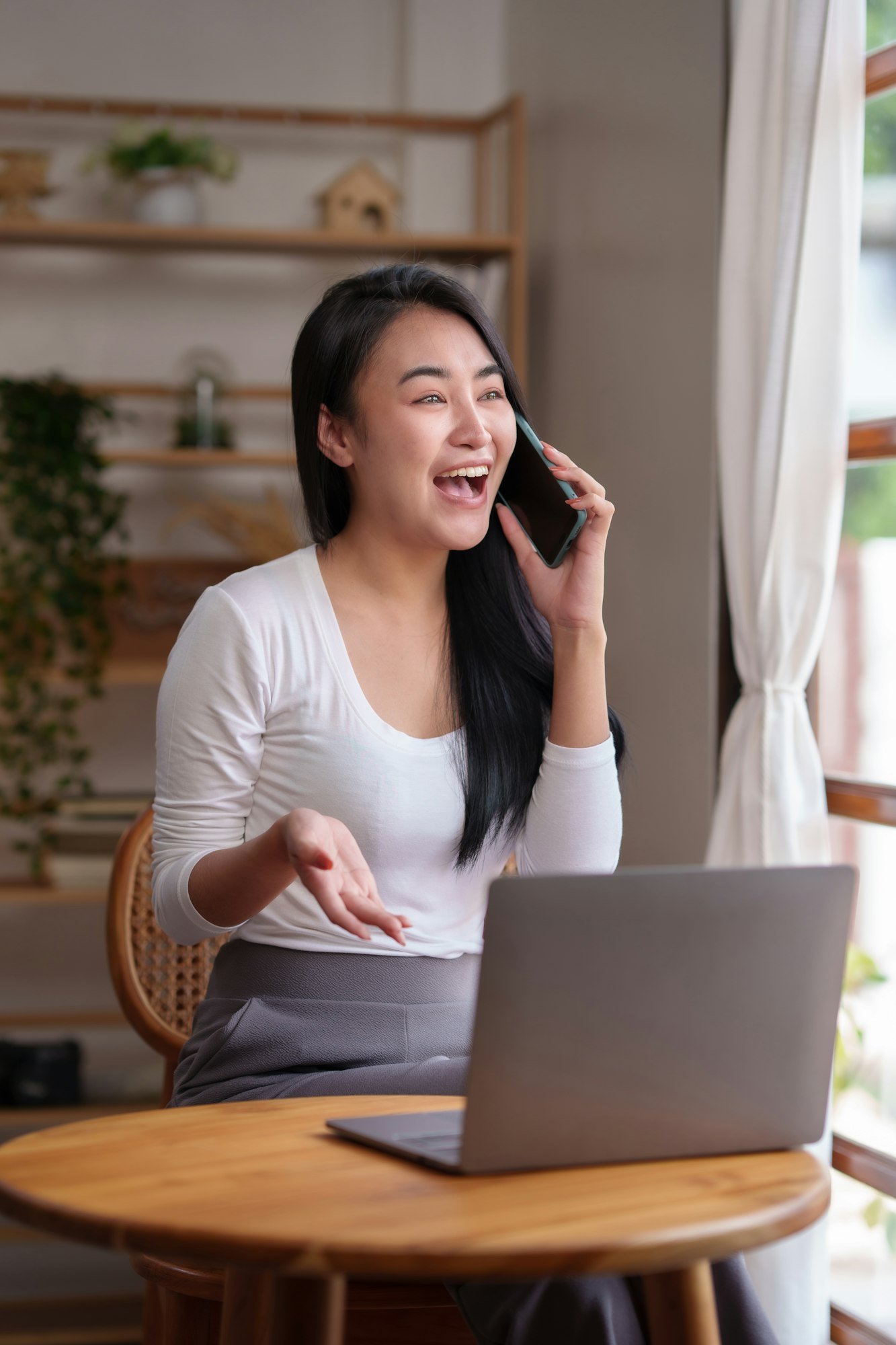 Asian woman sitting at home working on her laptop and talks on the phone comfortably and refreshed o